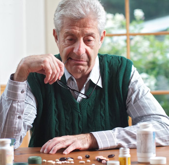 Senior man looking at many pills on a table Senior man with glasses in hand looks thoughtfully at many pills on table in front of him. Focus on man. Frontal view, square format, green and white color palette.